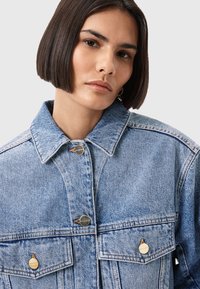 Giacca in denim azzurro chiaro con colletto classico, due tasche sul petto e bottoni in metallo argentato. Dettagli di cucitura lungo le cuciture e i bordi.