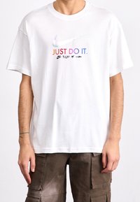 Vit bomullströja med en mångfärgad "JUST DO IT." grafik och Nike Swoosh-logotyp tryckt på framsidan. Korta ärmar, avslappnad passform.