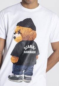 Grafická trička s medvědem v černém tričku s nápisem "GIANNI KAVANAGH". Bílé plátno ukazuje barevný design medvěda, džínové kalhoty a tenisky.