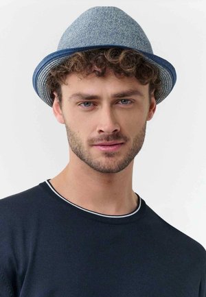 Junger Mann mit lockigem Haar, der einen blau-grauen Fedora-Hut und einen dunklen Pullover mit Rundhalsausschnitt vor einem hellen Hintergrund trägt.
