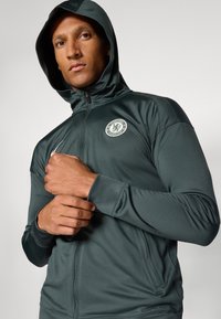 Chelsea FC hoodie in donkergroen, gemaakt van gladde stof, met een rits aan de voorkant, logo patch en aansluitende manchetten. Strakke, sportieve uitstraling.