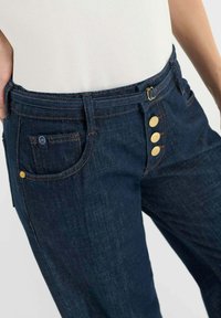 Donkerblauwe denim jeans met een klassiek ontwerp met vijf zakken, goude accenten op de knopen en contrasterende stiksels. Gladde textuur.
