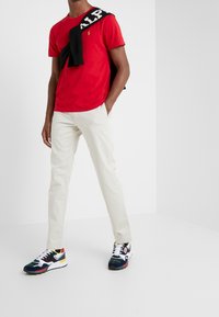 Rood T-shirt met korte mouwen, beige chino's en meerkleurige sportschoenen. Zwart trui over de schouders gedrapeerd met witte tekstdetails.