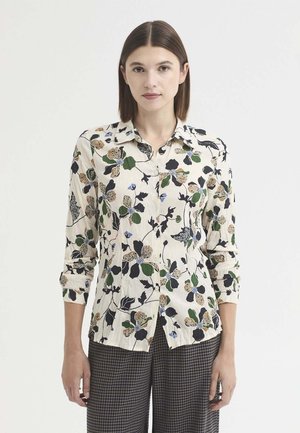 Femme aux cheveux bruns et raides porte un chemisier crème à imprimé floral vert, noir et marron et un pantalon à carreaux gris, debout sur un fond blanc.