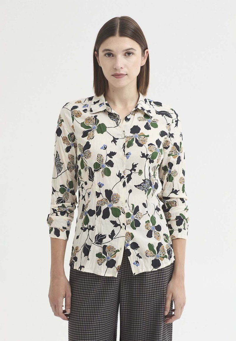 Femme aux cheveux bruns et raides porte un chemisier crème à imprimé floral vert, noir et marron et un pantalon à carreaux gris, debout sur un fond blanc.