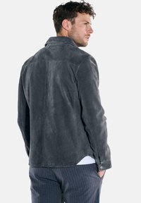 Graue Wildlederjacke mit Kragen, die eine glatte Textur aufweist. Markante Nähte entlang des Rückens und mit Knöpfen versehene Manschetten.