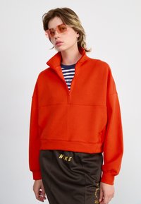 ORANSJE ZIPPER-FLEECE MED HØY KRAGE laget av mykt stoff, med cropped design og drop shoulders, kombinert med stilige mørkebrune shorts med gull aksenter.