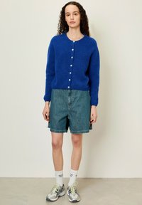 Blauwe fluffy cardigan met ronde hals en lichtblauwe knopen, gecombineerd met lichte denim shorts en sportieve schoenen. Gelaagde gebreide stof.
