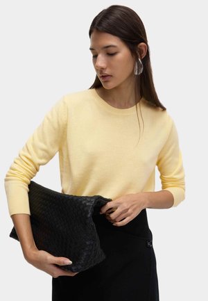 Maglione giallo di lana a maniche lunghe, abbinato a una pochette nera intrecciata e testurizzata. La modella tiene la pochette indossando una gonna nera.