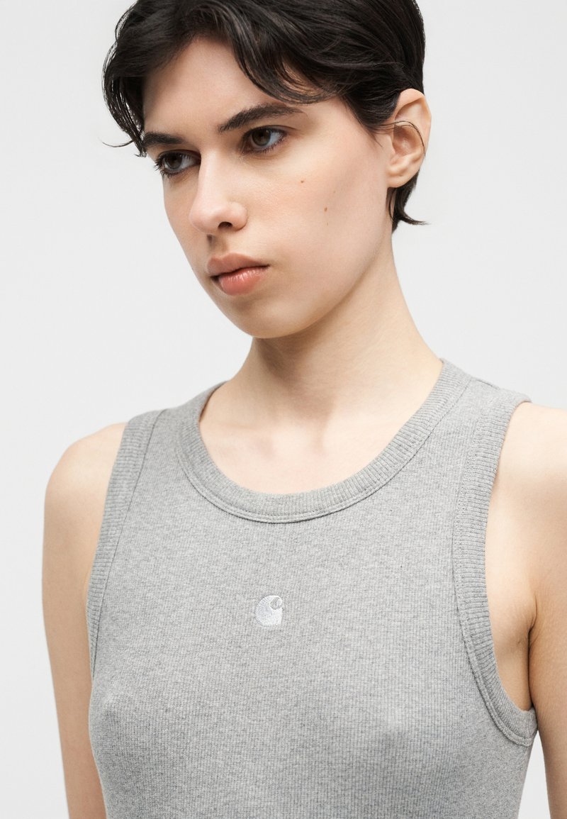 Jeune personne aux cheveux courts et foncés portant un débardeur gris clair à côtes, sans manches, sur un fond uni.