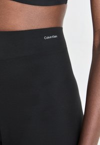 Detailný záber osoby v čiernych legínach s vysokým pásom, na ktorých je logo Calvin Klein na páse, pred svetlým pozadím.