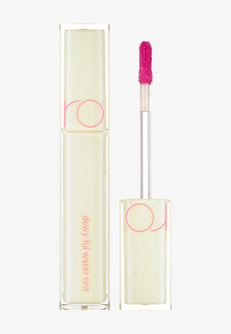 Tube rectangulaire clair avec une teinte jaune, des lettres roses et une wand applicateur rose. La formule semble liquide et éclatante.