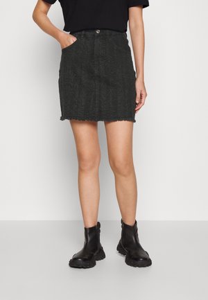 Kobieta nosząca czarną mini spódnicę z denimu w wzory, z postrzępionym dołem, czarne botki za kostkę oraz czarny krótki top z krótkim rękawem, ręka w kieszeni spódnicy.
