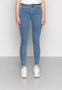 Calças skinny azuis, feitas de denim, com um corte alto, bolsos frontais e um ajuste justo até os tornozelos. Combinadas com sapatilhas brancas.