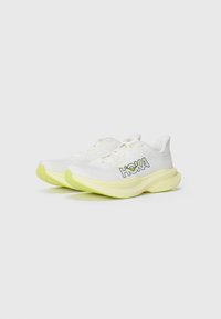 Des chaussures de course blanches avec une tige en maille texturée, des accents jaunes sur la semelle et un logo HOKA bien en vue sur le côté.