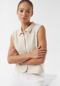 Beżowy dzianinowy top bez rękawów z kołnierzykiem, z guzikami z przodu i teksturowanym materiałem. Model z założonymi rękami, w jasnych spodniach.