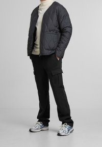 Vadderad svart jacka med dragkedja fram och en ficka, ihop med svarta cargobyxor och silverfärgad löparsneaker med blå detaljer.