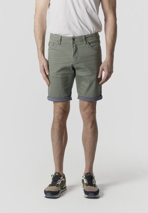 Mand iført olivengrønne shorts med opsmøg, hvid t-shirt og beige og sorte sneakers, stående mod en ensfarvet lys baggrund.