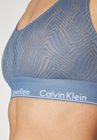 Blue mesh bralette with a zigzag pattern, featuring a wide elastic band with "Calvin Klein" logo in white. Soft, textured fabric.