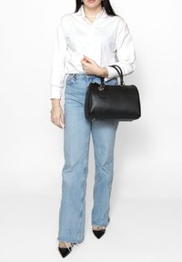 Zwarte leren handtas met een ronde vorm, gouden hardware accenten en een gestructureerde oppervlakte, vastgehouden door een persoon in een wit shirt en blauwe jeans.