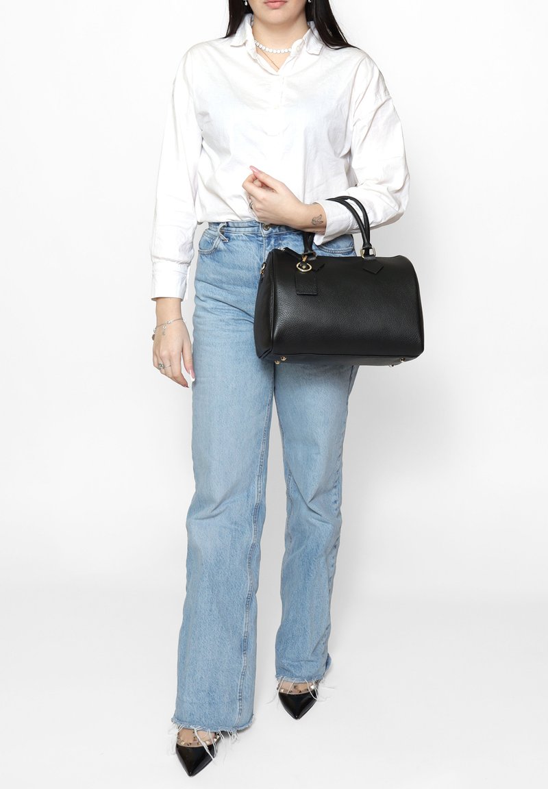 Zwarte leren handtas met een ronde vorm, gouden hardware accenten en een gestructureerde oppervlakte, vastgehouden door een persoon in een wit shirt en blauwe jeans.