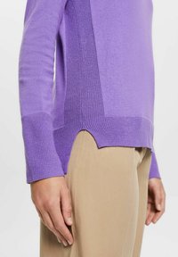 Lila stickad tröja med texturerade sidopaneler, ribbade manschetter och en hög-låg fåll. Bärs med ljusbeiga byxor som visar en figurnära silhuett.