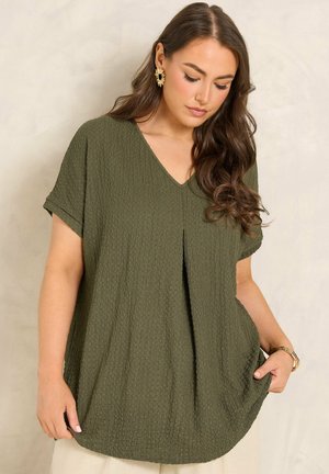 Femme aux longs cheveux bruns porte un haut texturé vert olive à col en V, des boucles d'oreilles anneaux dorées et des bracelets, debout devant un fond neutre.