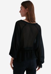 Czarna przezroczysta bluzka o luźnym kroju, z długimi, zwiewnymi rękawami i peplum na dole. Przezroczysta tekstura ukazuje warstwy pod spodem.