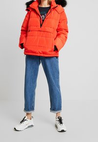 Röd vadderad jacka med huva av päls, utrustad med en framficka med dragkedja, kombinerad med löst sittande blå denimjeans och vita sneakers.