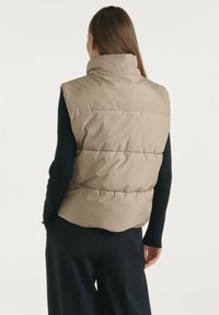 Beżowa kamizelka puchowa z wysokim kołnierzem i poziomym pikowaniem, pokazana od tyłu na jasnym tle, w zestawieniu z ciemnymi rękawami.