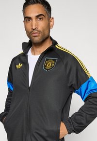 Melna rāvējslēdzēja jaka ar dzeltenām un zilām akcentēm, uz krūtīm redzams Mančestras United logo un apkaklē Adidas logo.