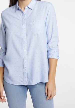 Vrouw die een lichtblauw overhemd met kleine blauwe stippen draagt en opgerolde mouwen, gecombineerd met lichtblauwe spijkerbroek.