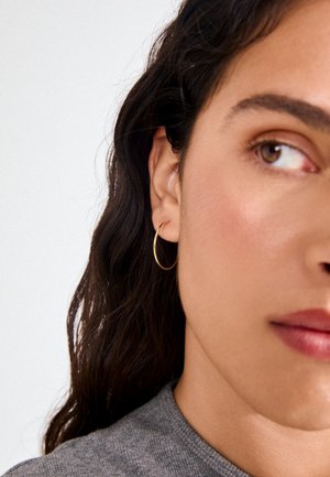 Gold hoop earring displayed in the ear; slender design with a smooth, shiny finish, complementary to the textured gray fabric of the shirt.