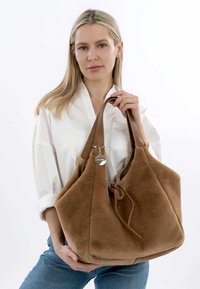 Sac à main en daim marron avec une texture douce, un dessus ouvert et une fermeture à cordon. Équipé de poignées en cuir et d'un accent en métal.