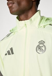 Lys grøn atletisk jakke lavet af glat, let materiale. Har grå Adidas-striber og et Real Madrid-emblem på brystet.