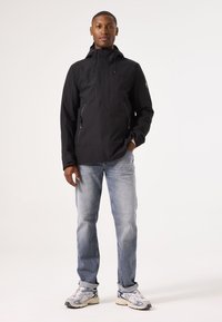 Black waterproof jacket with a hood, front zip pockets, elastic cuffs, paired with light blue jeans and grey sneakers.