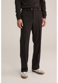 Mand iført mørkebrune skræddersyede bukser og hvide sneakers, stående på en ensartet beige baggrund.