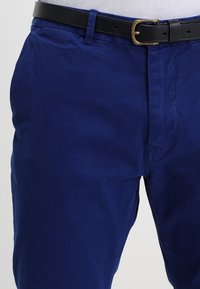 Donkerblauwe katoenen broek met een gladde textuur, voorzien van een zwarte riem met een gouden gesp. De broek heeft standaard zakken en een strak ontwerp.