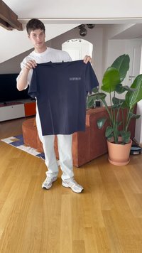T-shirt de algodão azul-marinho com gola redonda, mangas curtas e um pequeno gráfico na frente. Segurada por uma pessoa em um interior iluminado.