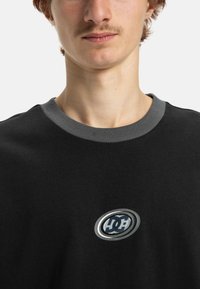 Sweatshirt noir avec un col rond gris et un logo ovale argenté avec des lettres noires et une étoile au centre. Texture douce, design décontracté.