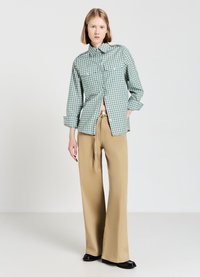 Chemise à carreaux verts et blancs à manches longues avec deux poches poitrine, associée à un pantalon beige à jambes larges et des ballerines noires.