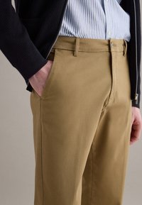 Hombre con pantalones beige, camisa de rayas azules y blancas metida por dentro, y suéter negro con cremallera, con la mano en el bolsillo.