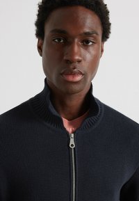 Maglione navy con zip, texture a costine e colletto alto, con zip argentata, indossato sopra una maglietta rosa.