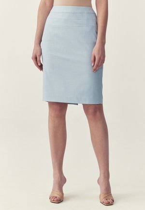 Kvinde iført en lyseblå knælængde blyantskørtd og beige åbne høje sandaletter, stående mod en ensfarvet baggrund.