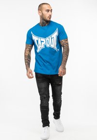 Mėlynas medvilnės marškinėliai su dideliu baltu "TAPOUT" logotipu, derinami su juodais suplyšusiais džinsais ir baltais sneakeriais. Modelio rankos puikuojasi tatuiruotėmis.