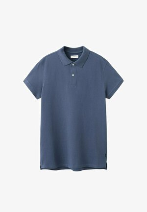 Junger Mann steht vor einer weißen Wand, trägt ein blaues Polohemd, hellblaue Jeans, braune Schuhe und einen dunklen Pullover, der über die Schultern gelegt ist.