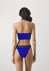 Mėlynas bikinių komplektas su bandeau tipo viršumi ir žemais šortais, turinčiais kontrastingą juodą juostą. Lygus, blizgus paviršius su minimaliais priedais.