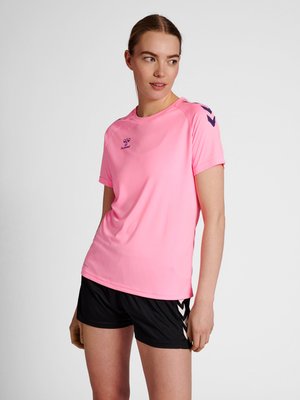 Vrouw draagt een roze Hummel sport T-shirt en zwarte korte broek, staat met ontspannen armen en kijkt iets opzij tegen een effen achtergrond.