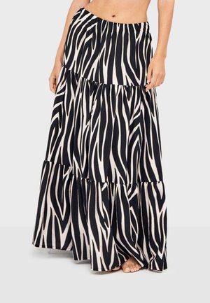 Mujer con falda larga en capas con estampado de cebra en blanco y negro, de pie descalza contra un fondo claro y liso.