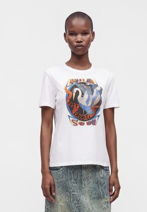 Persona joven con cabello corto que lleva una camiseta blanca con un gráfico colorido de un pájaro "Wild Soul" y vaqueros azul claro con estampado.